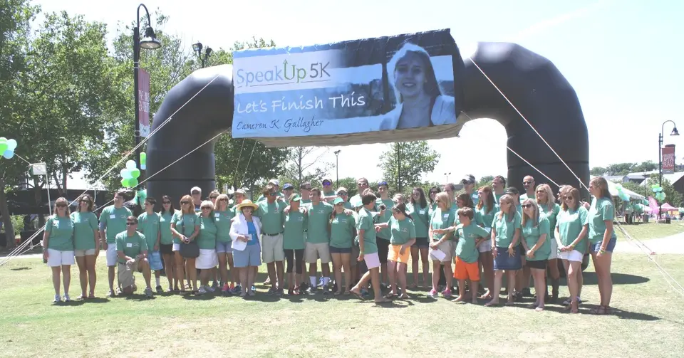 Group in green t-shirts stands in front of a black inflatable arch with a SpeakUp 5K banner reading Let's Finish This