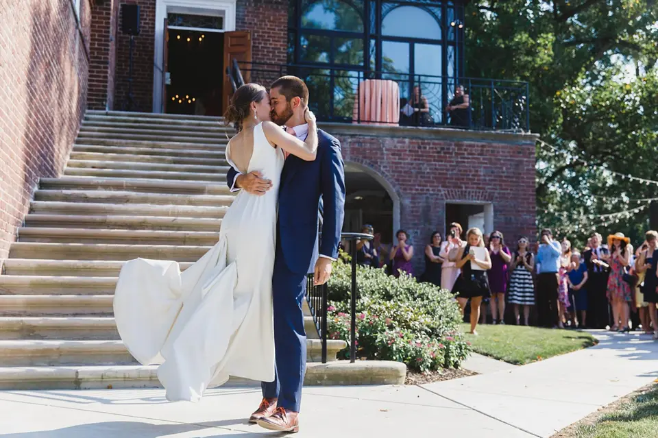Groom lifting the bride in a kiss on stone steps while guests watch from a lawn