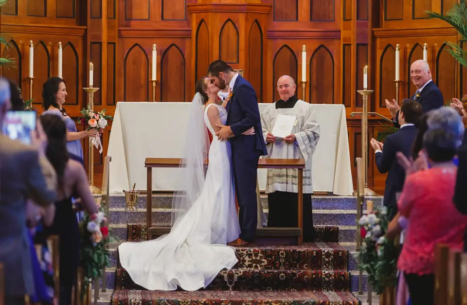 Bride and groom kissing at a church altar as a priest and guests look on
