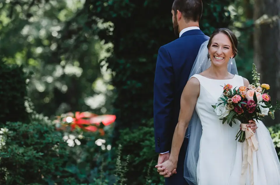 Bride smiling with a colorful bouquet, holding the groom's hand as he faces away in a navy suit