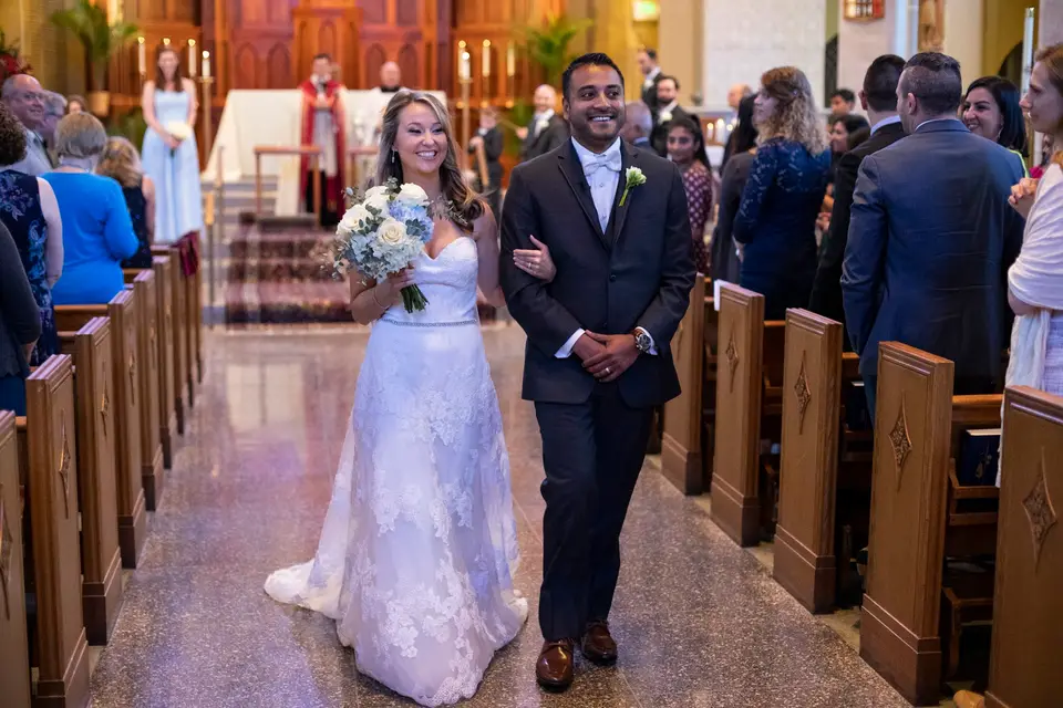 Atit and Maura walking up the aisle of a church past guests in the pews