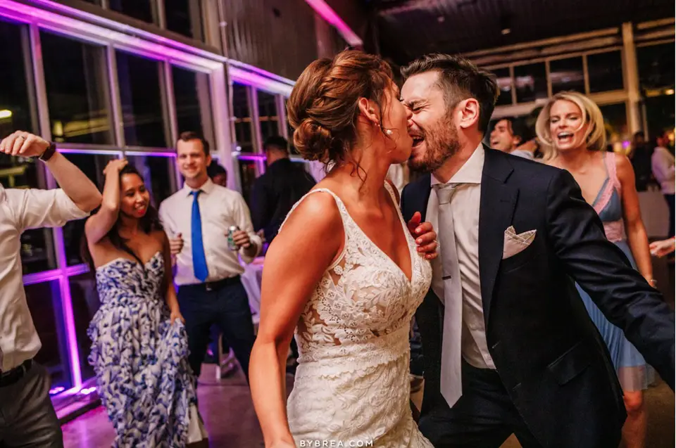 Bride and groom kissing and laughing on the dance floor under purple lighting with guests behind
