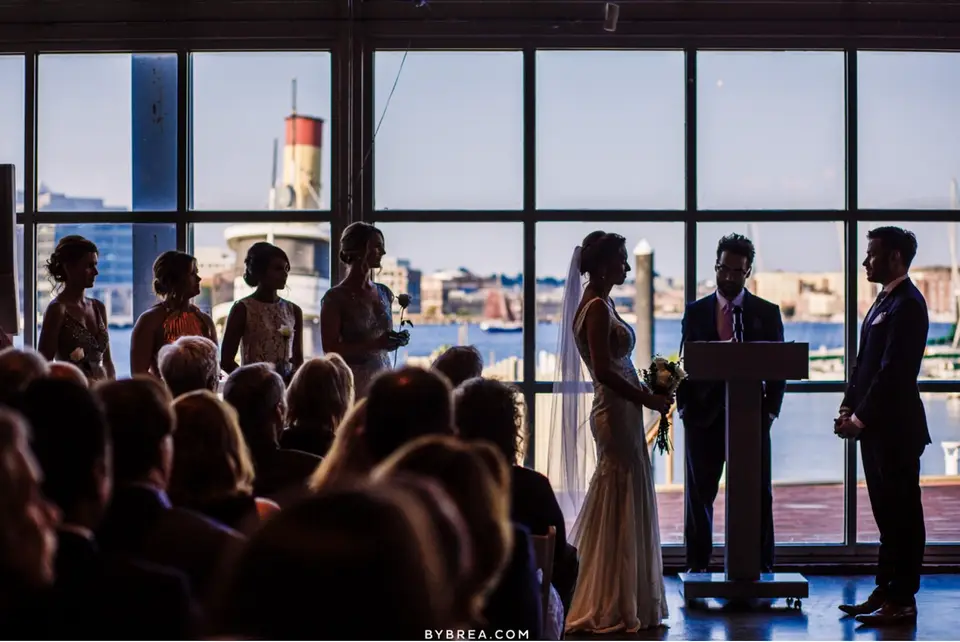 Bride and groom at the altar in front of waterfront windows as wedding party and guests look on