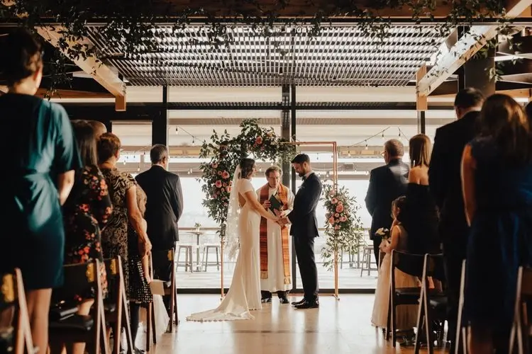 Couple facing an officiant during a ceremony with a floral arch and floor-to-ceiling windows behind them