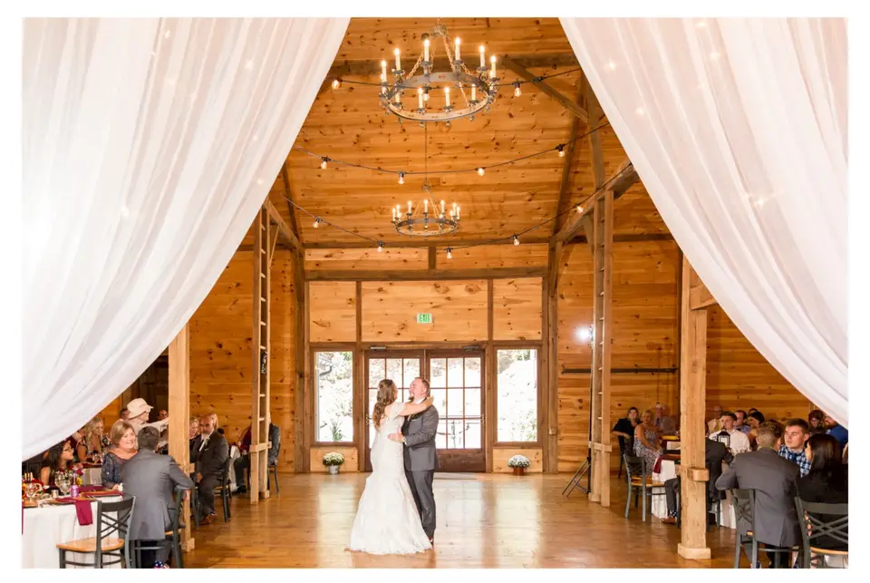 Newlyweds share a first dance in a timber barn with white draping and chandeliers while guests watch
