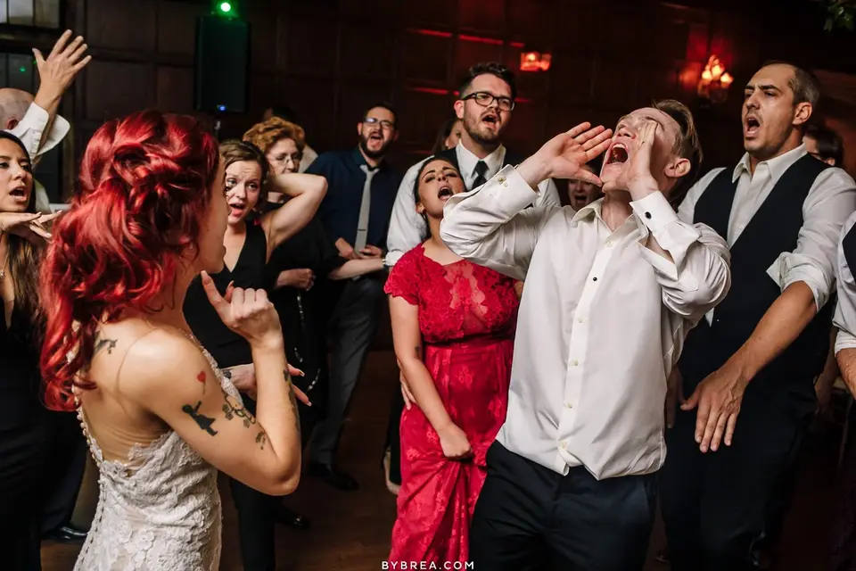 Guests with heads tilted back singing and cheering on the dance floor, a tattooed bride at left
