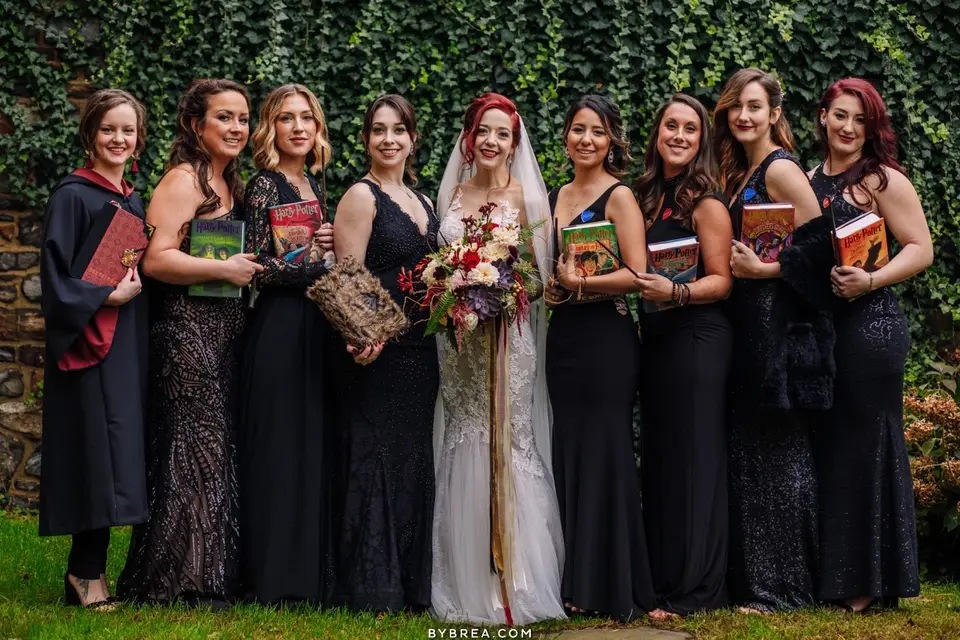 Bride holding a bouquet with seven bridesmaids in black dresses holding books against an ivy wall