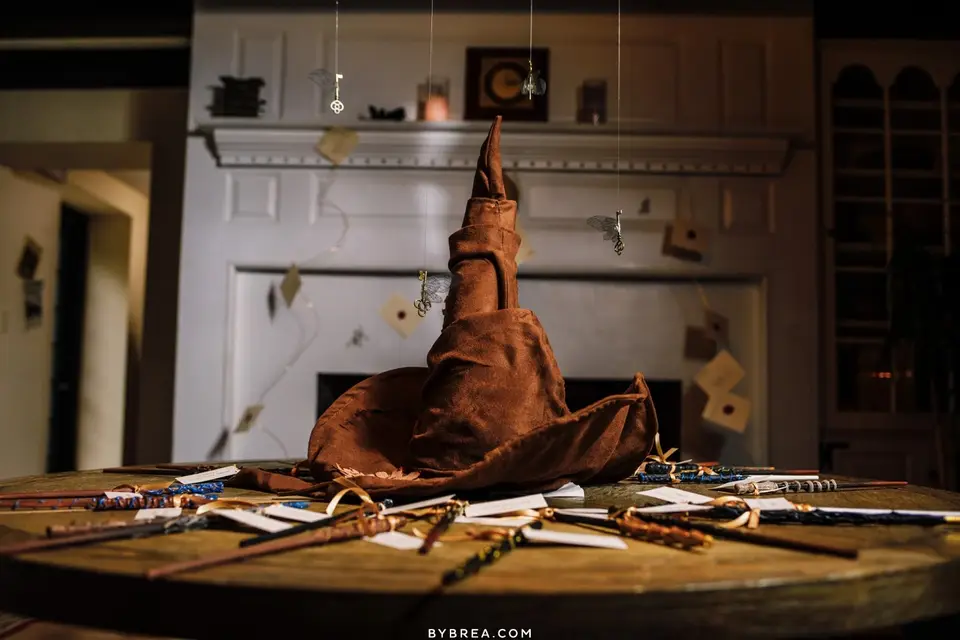 Brown wizard hat on a wooden table with wands and hanging ornaments in front of a fireplace