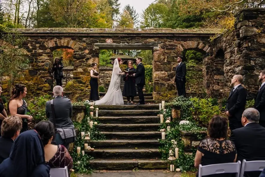 Outdoor wedding ceremony on stone steps among ruins as guests watch and an officiant reads