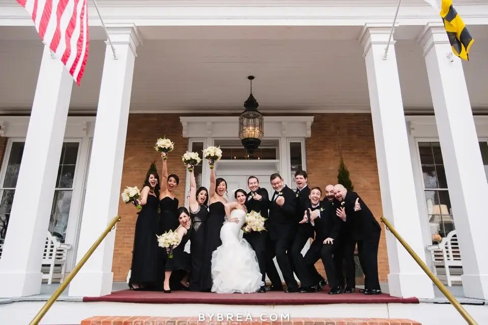 Groomsmen, bridesmaids in black and the bride cheering on a columned porch with an American flag