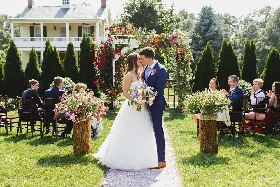 Couple kissing at the end of an outdoor aisle in front of a floral arch, with guests and a house behind