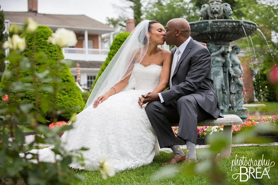 Bride and groom kissing on a stone bench beside a fountain in a garden