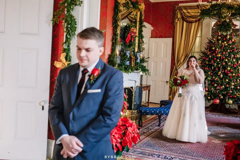 Groom in a navy suit faces away while the bride waits behind him in a room with a Christmas tree