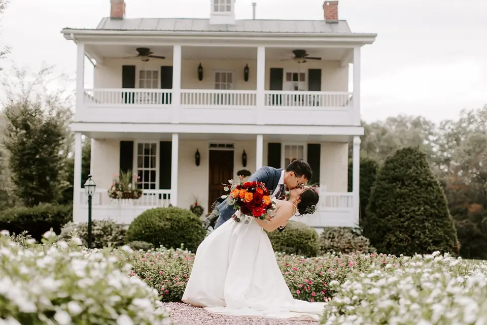 Groom dips the bride for a kiss in front of a two-story white house, holding an orange bouquet