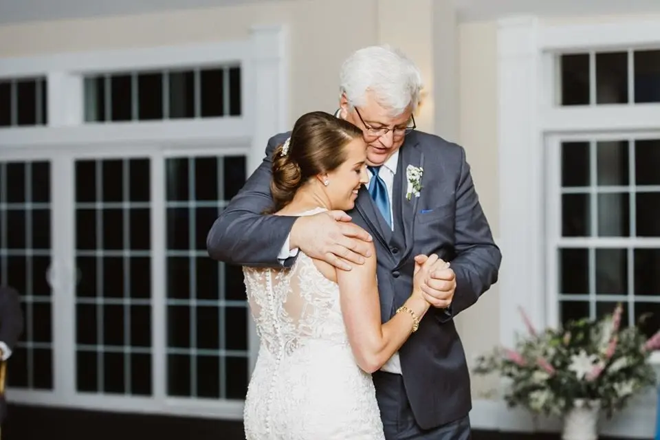 Bride dancing with her father, who wears a gray suit, in front of large windows