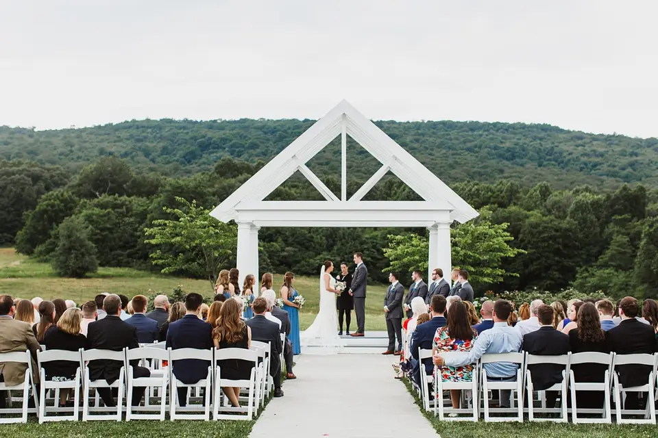 Outdoor ceremony at a white pavilion with guests seated in rows and wooded mountains behind