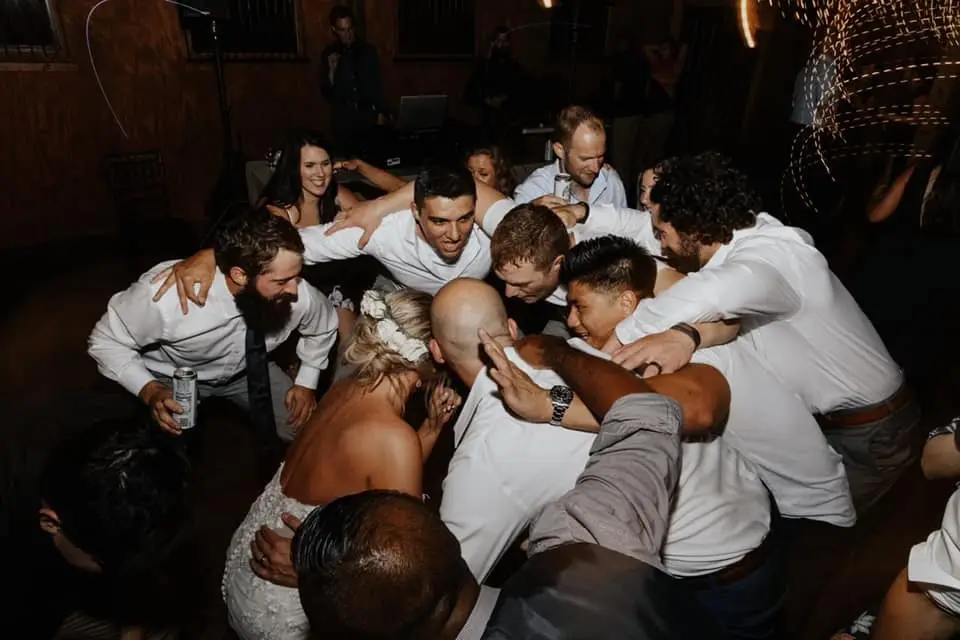Groomsmen and guests huddled in a tight circle on the dance floor around the bride at a barn reception