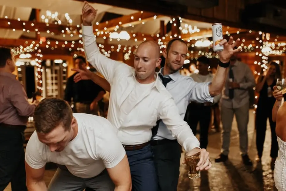 Groomsmen dancing and raising drinks and a beer can at a barn reception under string lights