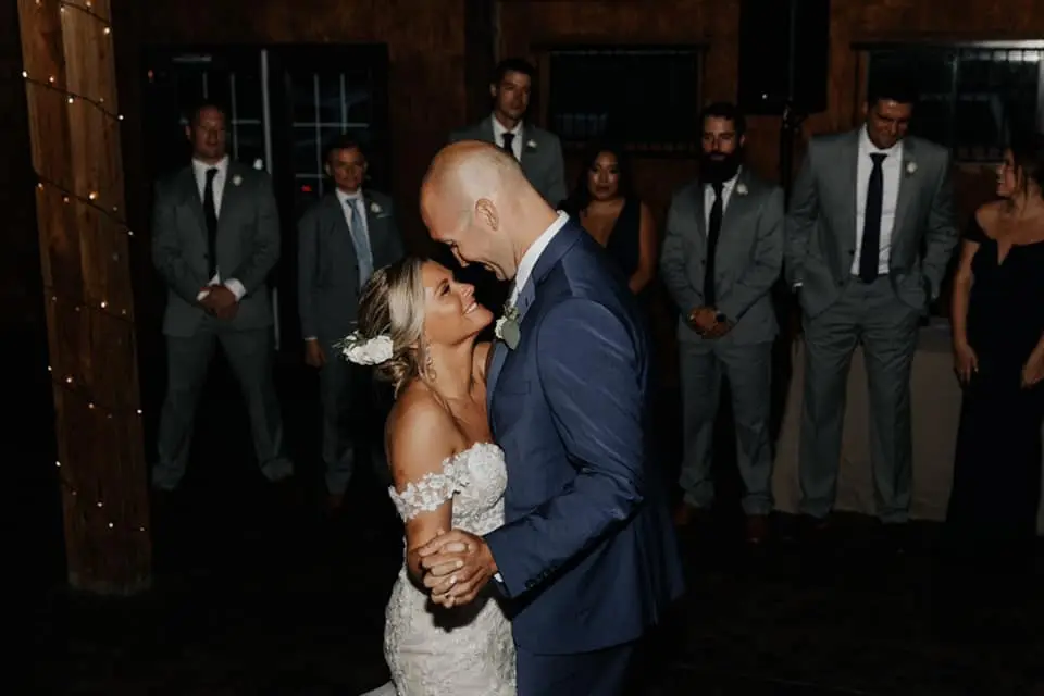 Bride and groom sharing a first dance in a barn as groomsmen watch from behind