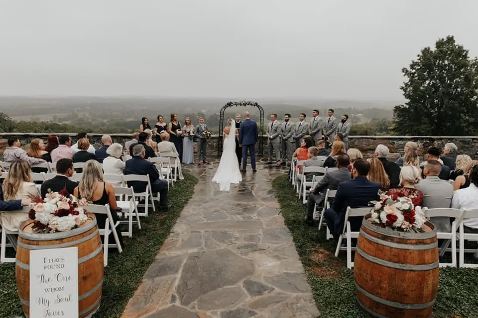 Outdoor wedding ceremony on a hilltop at a stone patio aisle, with guests seated and a barrel at each side