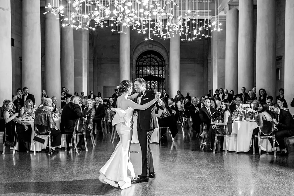 Black and white photo of a bride and groom dancing in a grand columned hall with chandeliers