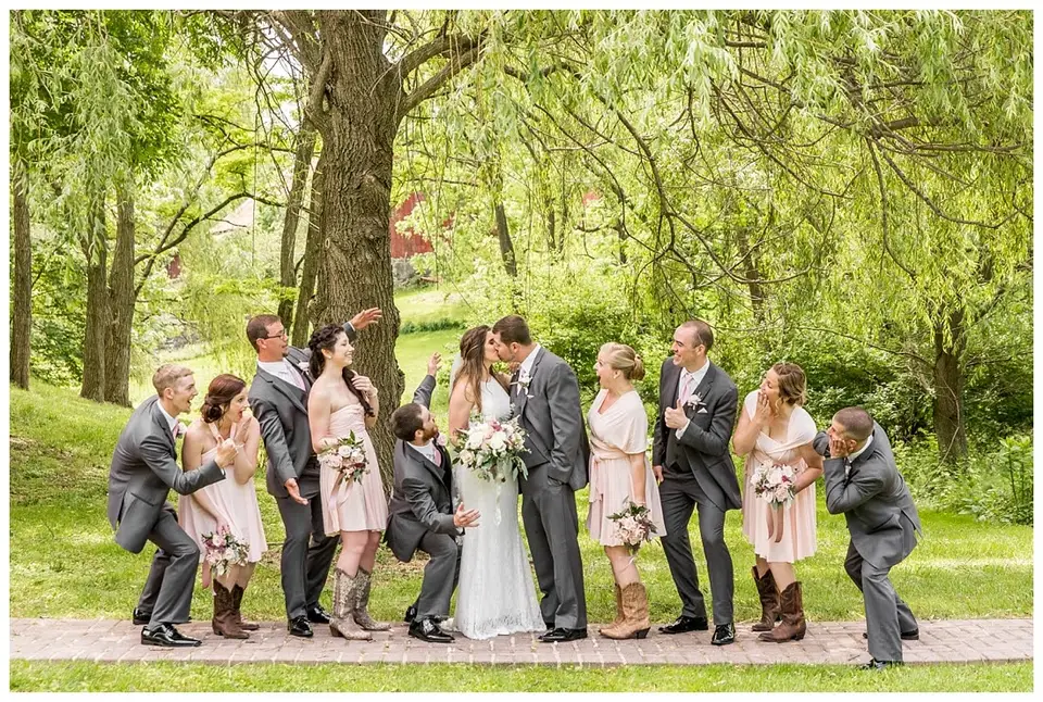 Bride and groom kiss under a willow tree while bridesmaids and groomsmen cheer and pose around them