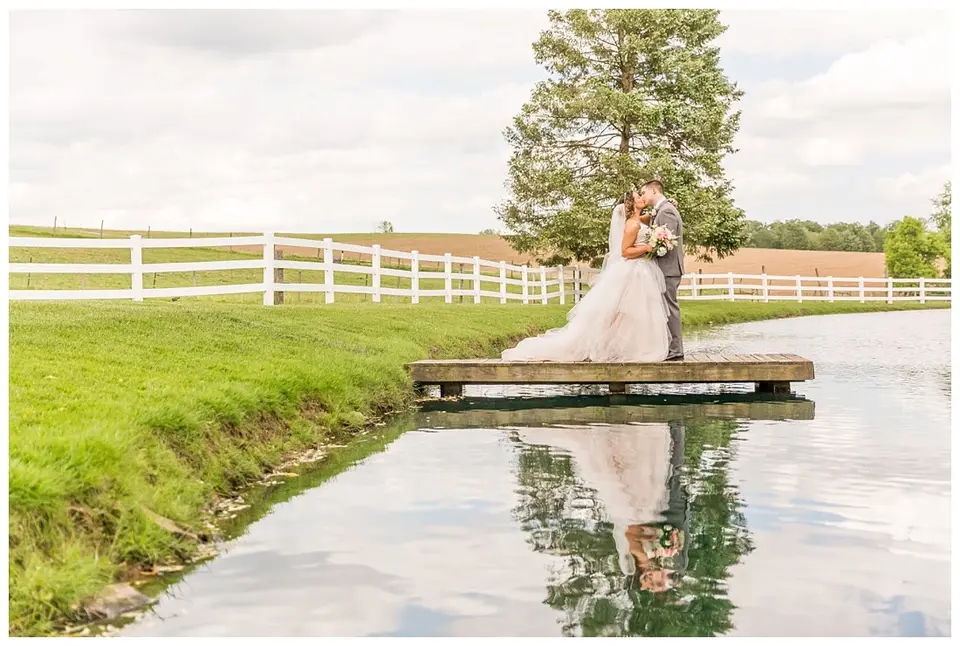 Bride and groom kiss on a small dock over a pond with a white fence and tall pine tree behind them