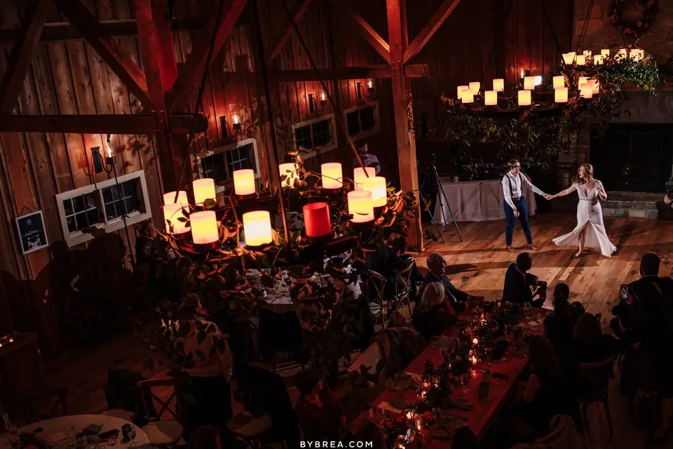 Overhead view of a bride and groom dancing in a rustic barn with glowing lanterns and tables below