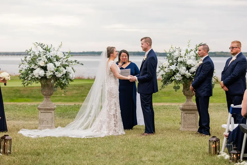 Bride and groom exchanging vows at a waterfront ceremony with large white floral arrangements