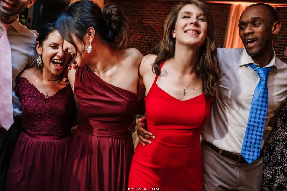 Group of women and a man in red and burgundy dresses laughing with arms around each other at a reception