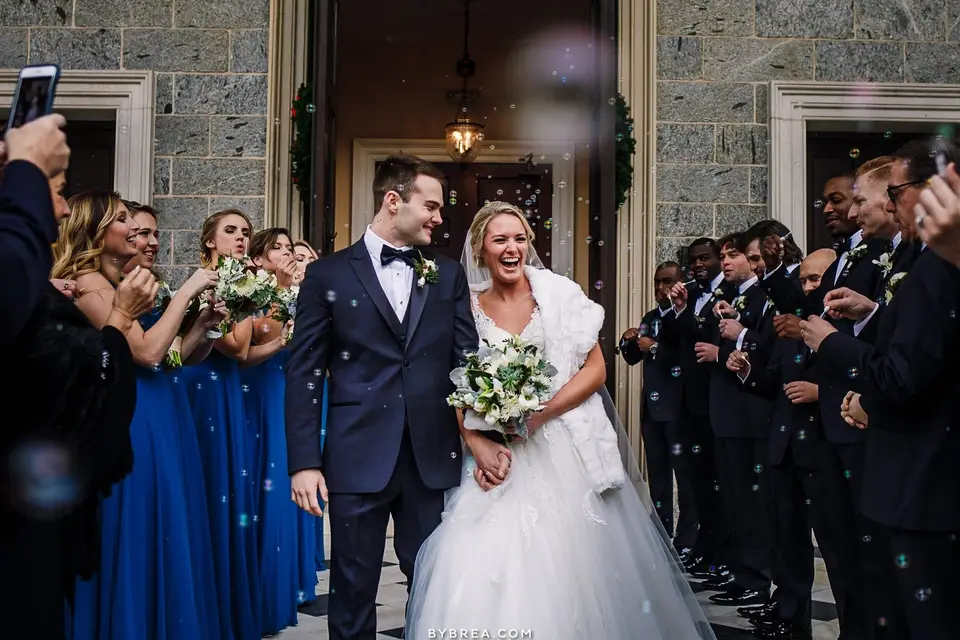 Bride and groom laughing as they exit a stone building past guests blowing bubbles