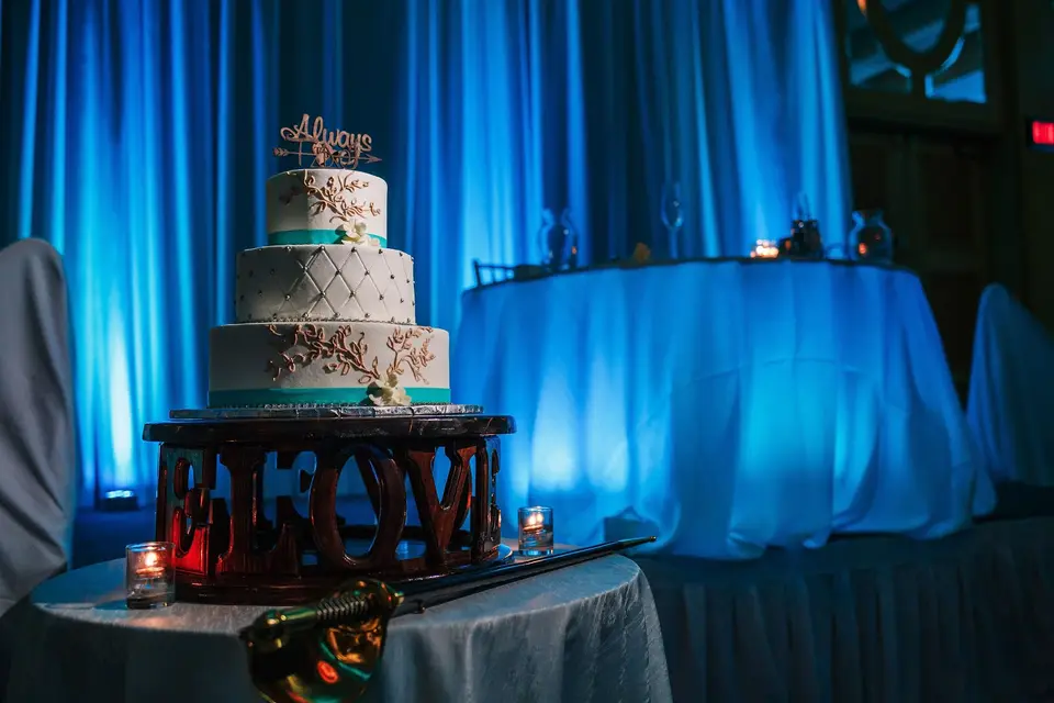 Three-tier wedding cake with a sword on a table in front of blue uplit drapes