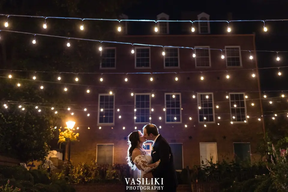 Bride and groom kissing at night in front of a brick building strung with cafe lights