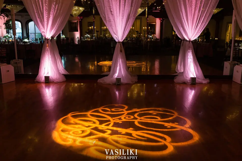 Gold monogram light projected on a wooden dance floor in front of pink-lit draped columns