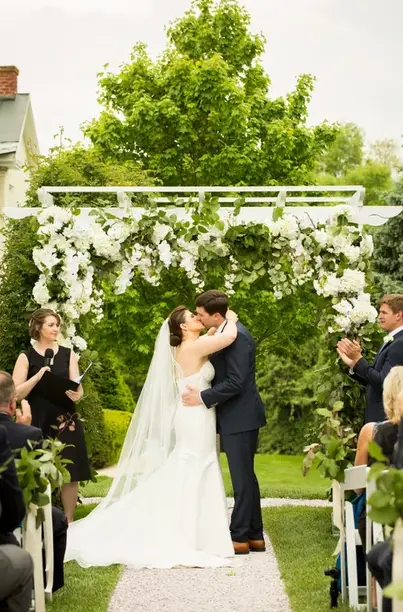 Bride and groom kissing under a floral arch on a lawn while an officiant and guests look on