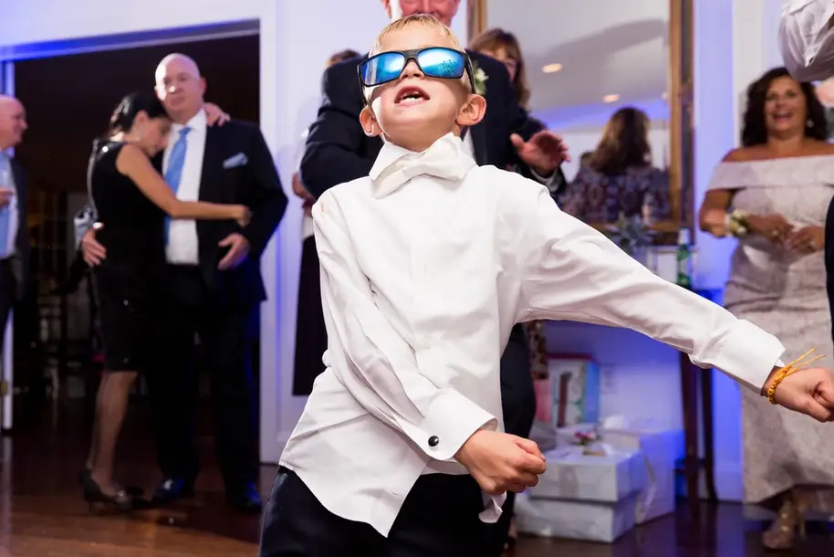 Young boy in a white tuxedo shirt and blue sunglasses dancing at a wedding reception