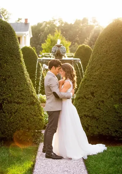 Bride and groom embracing in a formal garden between trimmed hedges with a fountain behind them