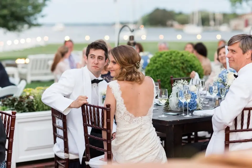 Bride in a lace gown seated at a reception table with groom and guests beside a waterfront lawn