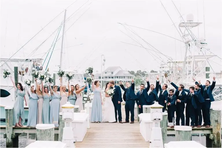 Wedding party on a marina dock cheering with raised arms as the couple kisses, bridesmaids in light blue