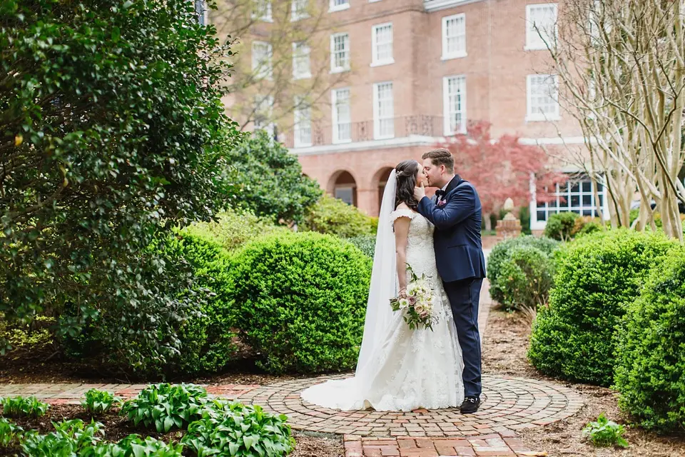Bride and groom kissing on a brick garden path lined with trimmed shrubs and a brick building behind