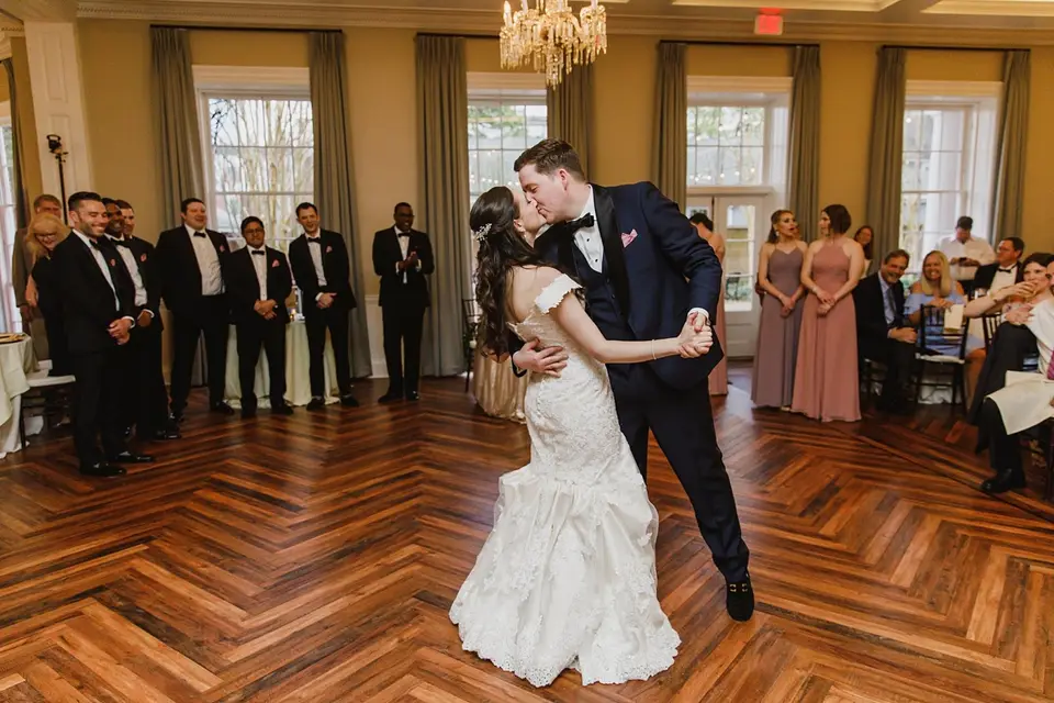 Bride and groom kissing during their first dance on a herringbone wood floor with wedding party watching