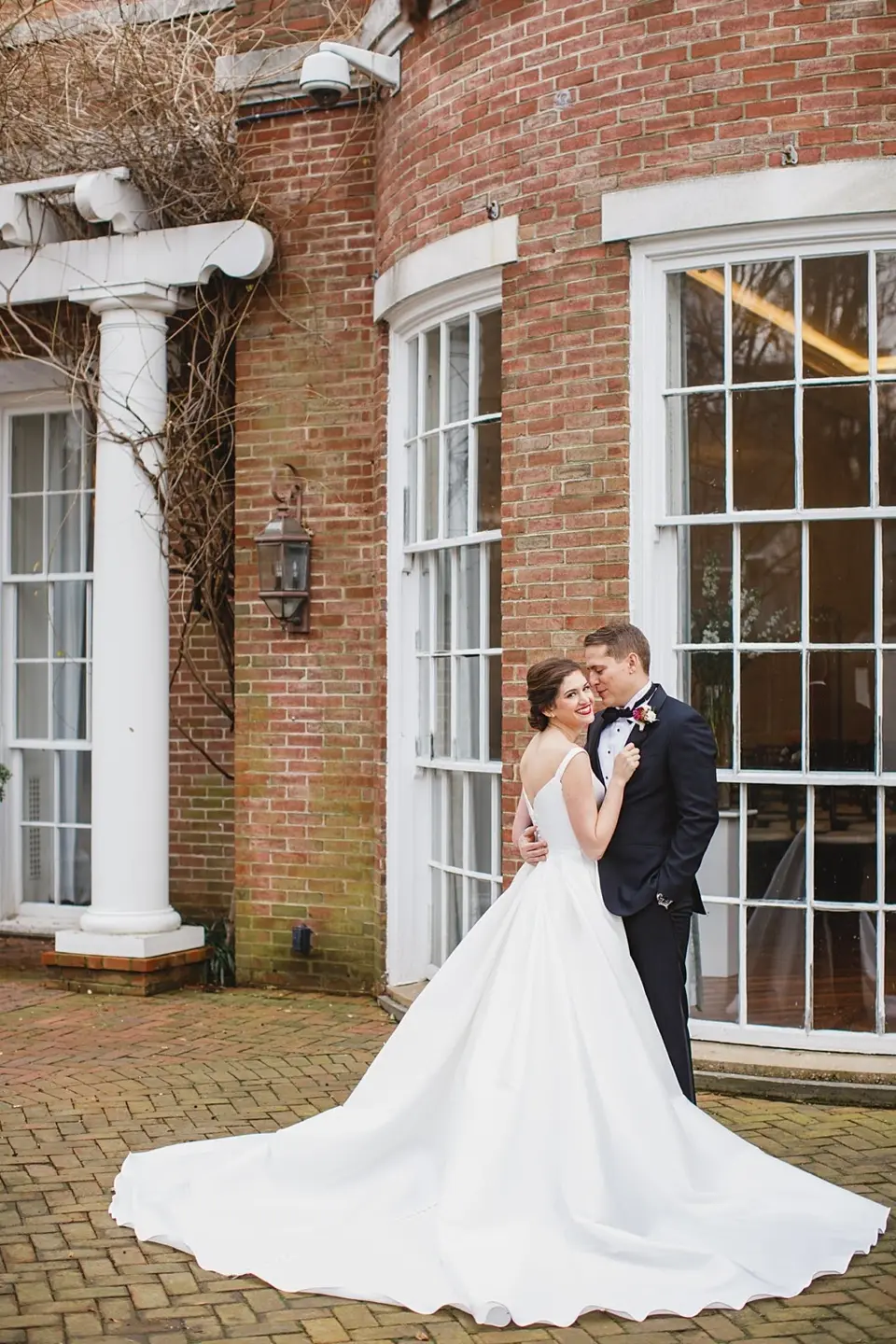 Bride in a long white gown with a train embraced by the groom beside a brick building with tall windows