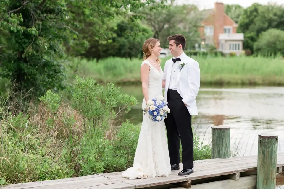 Bride and groom in a white dinner jacket standing hand in hand on a wooden dock beside a creek