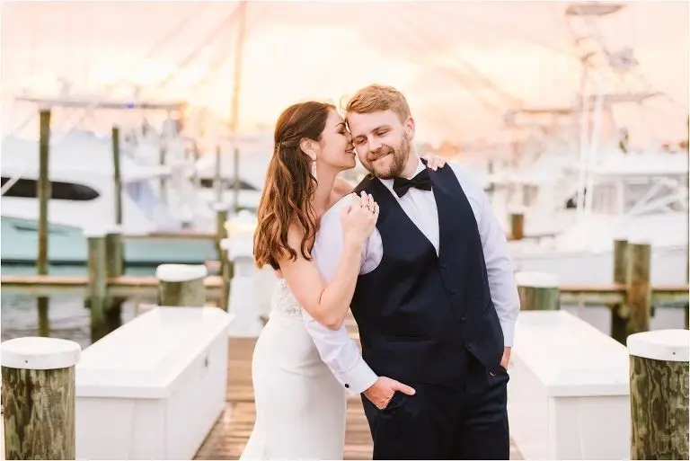 Groom embracing the bride from behind on a marina dock at sunset