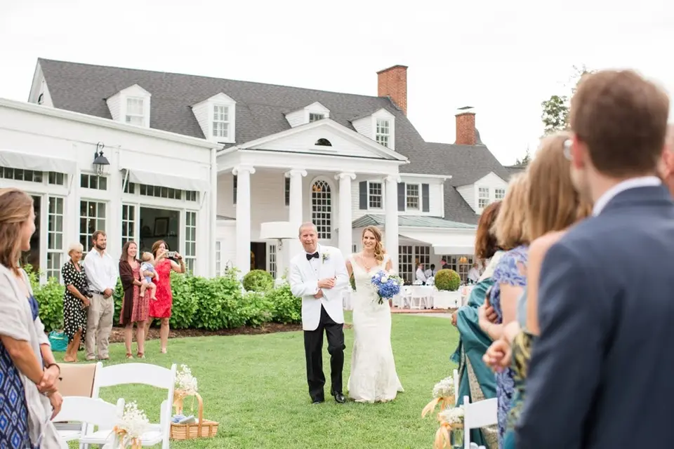 Bride on her father's arm walking across a lawn toward guests with a white waterfront inn behind them