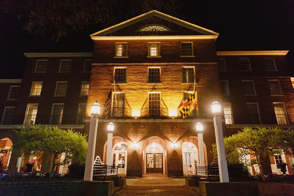 Historic brick inn lit by warm lights at night with Maryland and American flags and white columns