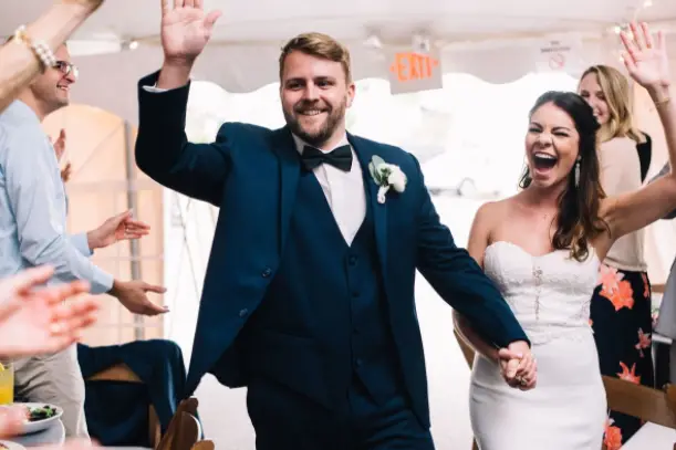 Groom in a navy vest and bow tie and laughing bride walking into a tent reception with guests cheering