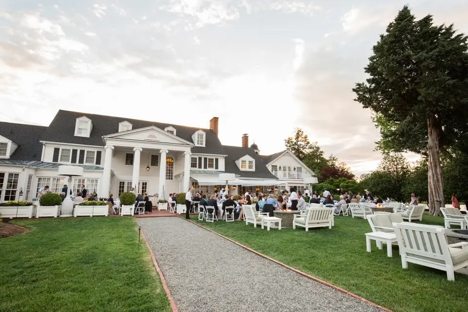 Guests gathered on a lawn and patio with white chairs outside a white waterfront inn at dusk