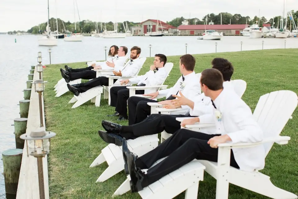 Groomsmen in white dinner jackets reclining in white Adirondack chairs on a waterfront lawn