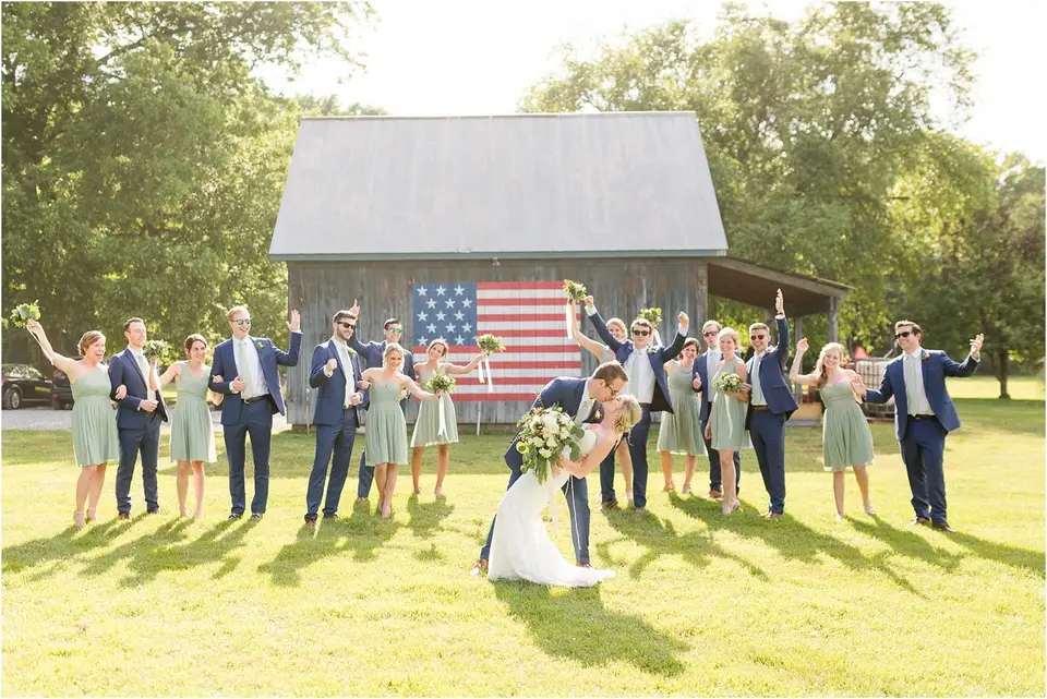 Wedding party cheering with raised arms around the kissing couple in front of a barn with an American flag